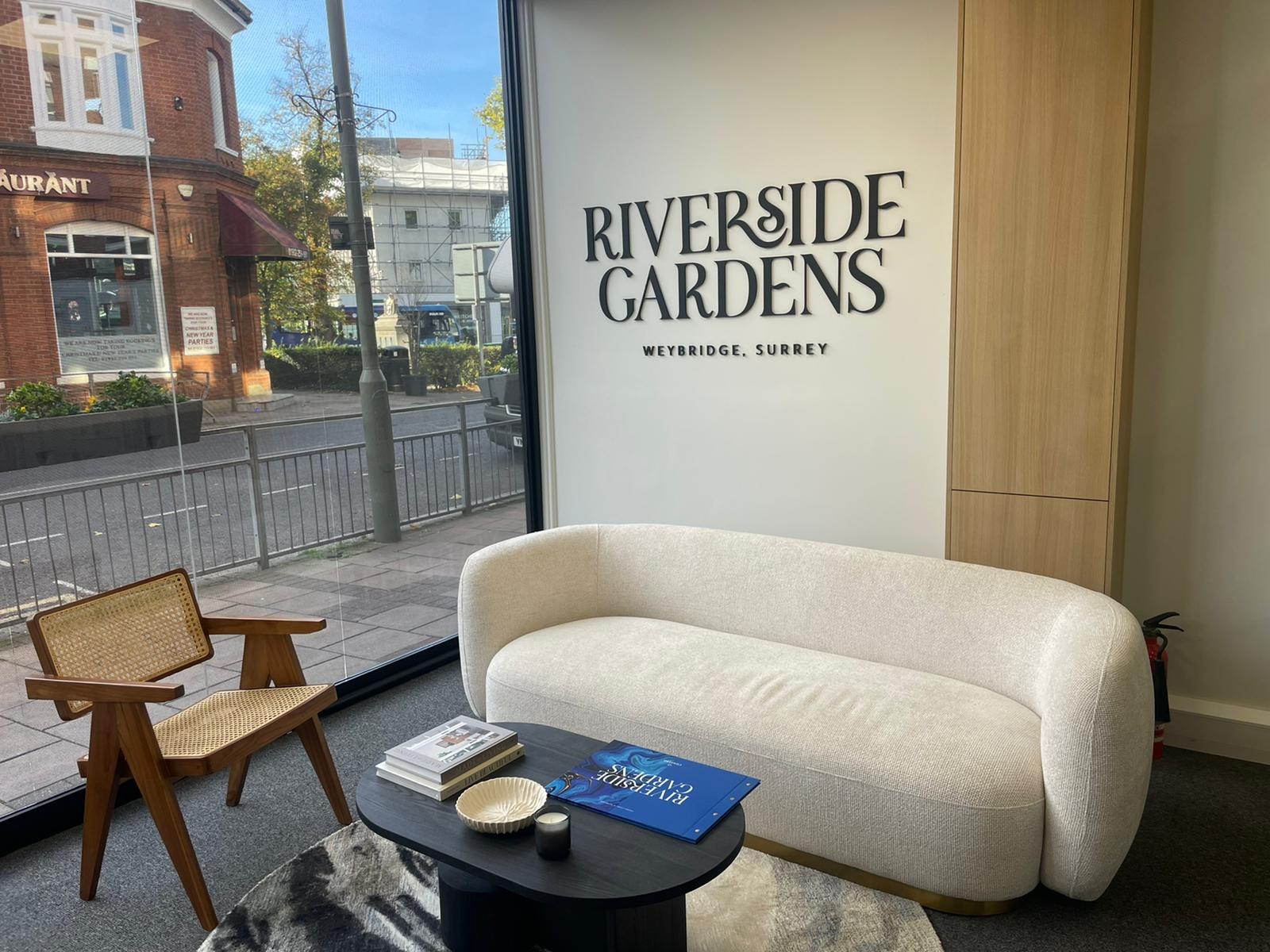 Riverside Gardens marketing suite with the development name in dimensional lettering, a curved bouclé sofa and full-height glazing to the street, Weybridge — design and styling by HDEY